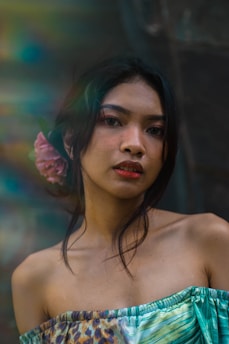 A young woman with long dark hair adorned with a pink flower, wearing an off-the-shoulder top with a colorful pattern. She is looking directly at the camera with a calm expression, set against a blurred and softly lit background with rainbow-like lens flares.