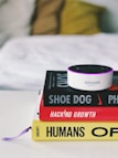 A small Amazon Echo device is placed on top of a stack of three books. The titles visible are 'Shoe Dog', 'Hacking Growth', and 'Humans', each with distinctive colors and fonts. The books are neatly stacked on a white surface, and in the background, a blurred, cozy-looking white and yellow pillow or blanket is visible.
