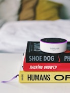 A small Amazon Echo device is placed on top of a stack of three books. The titles visible are 'Shoe Dog', 'Hacking Growth', and 'Humans', each with distinctive colors and fonts. The books are neatly stacked on a white surface, and in the background, a blurred, cozy-looking white and yellow pillow or blanket is visible.