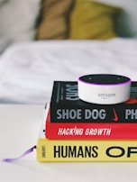 A small Amazon Echo device is placed on top of a stack of three books. The titles visible are 'Shoe Dog', 'Hacking Growth', and 'Humans', each with distinctive colors and fonts. The books are neatly stacked on a white surface, and in the background, a blurred, cozy-looking white and yellow pillow or blanket is visible.