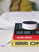 A small Amazon Echo device is placed on top of a stack of three books. The titles visible are 'Shoe Dog', 'Hacking Growth', and 'Humans', each with distinctive colors and fonts. The books are neatly stacked on a white surface, and in the background, a blurred, cozy-looking white and yellow pillow or blanket is visible.