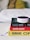 A small Amazon Echo device is placed on top of a stack of three books. The titles visible are 'Shoe Dog', 'Hacking Growth', and 'Humans', each with distinctive colors and fonts. The books are neatly stacked on a white surface, and in the background, a blurred, cozy-looking white and yellow pillow or blanket is visible.