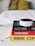 A small Amazon Echo device is placed on top of a stack of three books. The titles visible are 'Shoe Dog', 'Hacking Growth', and 'Humans', each with distinctive colors and fonts. The books are neatly stacked on a white surface, and in the background, a blurred, cozy-looking white and yellow pillow or blanket is visible.