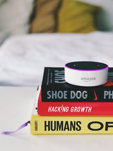 A small Amazon Echo device is placed on top of a stack of three books. The titles visible are 'Shoe Dog', 'Hacking Growth', and 'Humans', each with distinctive colors and fonts. The books are neatly stacked on a white surface, and in the background, a blurred, cozy-looking white and yellow pillow or blanket is visible.