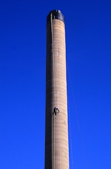 A tall, cylindrical industrial chimney rises against a clear blue sky. A person is climbing the structure, using ropes and harness equipment, midway up the chimney.