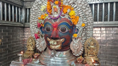 A large, ornate deity head crafted from metal with intricate details and colorful paint, adorned with garlands of fresh marigold and other flowers. It is placed on a pedestal flanked by two smaller metal statues, with a small oil lamp lit at the side. The background features metallic brick-patterned walls.