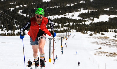 A person wearing a green helmet, sunglasses, a red sleeveless top, and blue shorts is skiing uphill on a snowy slope. Other skiers are visible in the background, making their way down the piste. The scene is set in a mountainous area with snow-covered trees in the distance.
