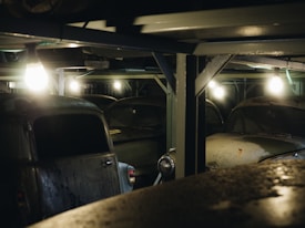 Dimly lit, dusty vintage cars are parked under a metal structure, with several lights illuminating the area. The scene appears to be a garage or storage space for classic vehicles, with a focus on the neglected and aged condition of the cars.