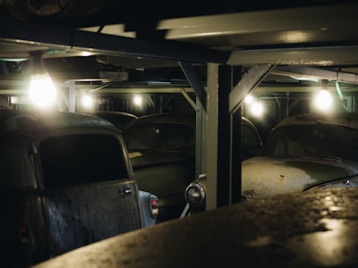 Dimly lit, dusty vintage cars are parked under a metal structure, with several lights illuminating the area. The scene appears to be a garage or storage space for classic vehicles, with a focus on the neglected and aged condition of the cars.