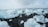 Large icebergs of varying sizes and shapes are scattered across a black sand beach. A person stands among the ice, gazing at the scene, with a camera tripod beside them. The sky is overcast, adding a dramatic effect to the landscape.