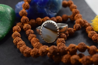 A close-up view of a silver ring with a striped stone centerpiece placed among a string of rudraksha beads. The background features a green stone and a blue rock, adding to the earthy and spiritual ambiance.