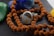 A close-up view of a silver ring with a striped stone centerpiece placed among a string of rudraksha beads. The background features a green stone and a blue rock, adding to the earthy and spiritual ambiance.