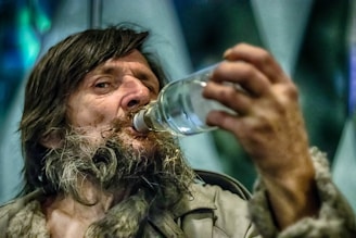 A person with long hair and a rugged beard is drinking from a clear bottle. The expression on their face shows focus or concentration. The background is blurred, with hints of blue and green tones.