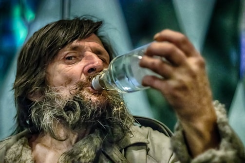A person with long hair and a rugged beard is drinking from a clear bottle. The expression on their face shows focus or concentration. The background is blurred, with hints of blue and green tones.