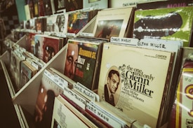 A collection of vinyl records is neatly arranged in a music store display. Various album covers are visible, featuring vintage designs and famous musicians like Glenn Miller, with organizer tabs labeling different artists.
