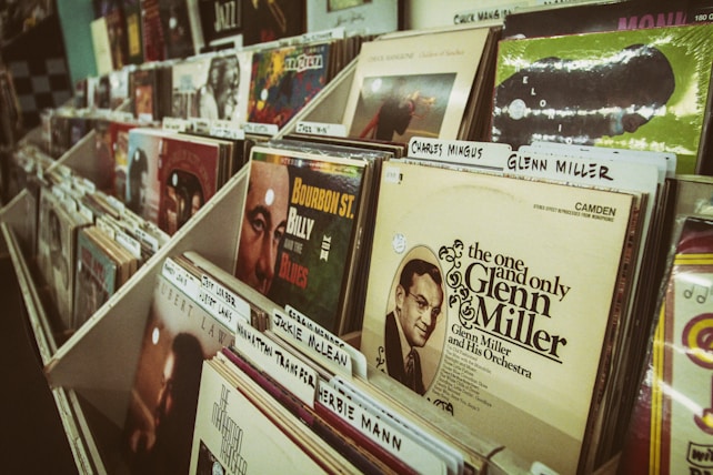 A collection of vinyl records is neatly arranged in a music store display. Various album covers are visible, featuring vintage designs and famous musicians like Glenn Miller, with organizer tabs labeling different artists.