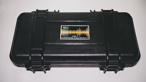 A black, hard-shell case with reinforced edges and a label in the center featuring sound wave graphics and the text 'DEITY S-Mic 2, Broadcast Quality Shotgun Microphone'. The case is sitting on a white surface.