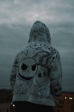 A person is standing with their back to the camera, wearing a hoodie covered in intricate, gothic-style drawings. The designs include various skull motifs, graveyard imagery, and stylized text. The sky is overcast, creating a moody atmosphere.