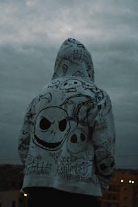 A person is standing with their back to the camera, wearing a hoodie covered in intricate, gothic-style drawings. The designs include various skull motifs, graveyard imagery, and stylized text. The sky is overcast, creating a moody atmosphere.