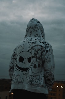 A person is standing with their back to the camera, wearing a hoodie covered in intricate, gothic-style drawings. The designs include various skull motifs, graveyard imagery, and stylized text. The sky is overcast, creating a moody atmosphere.