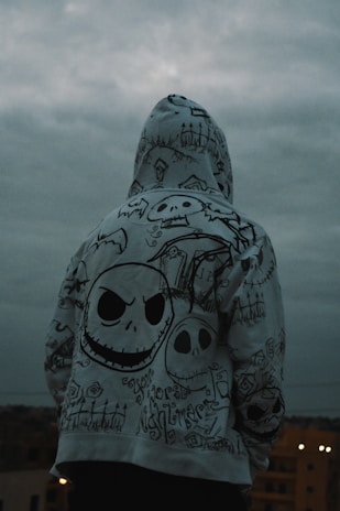 A person is standing with their back to the camera, wearing a hoodie covered in intricate, gothic-style drawings. The designs include various skull motifs, graveyard imagery, and stylized text. The sky is overcast, creating a moody atmosphere.