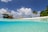 A tropical beach scene with a thatched-roof bungalow on stilts over turquoise water. Palm trees are on a sandy beach under a blue sky with fluffy white clouds. The view is partially submerged, showing clear water with ripples.