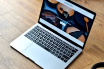 A sleek laptop with a silver body is open, displaying a webpage featuring wooden products. The wood-themed website includes images of handcrafted wooden items and text promoting their uniqueness and premium quality. The laptop rests on a wooden floor, complementing the aesthetic of the website displayed.