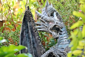 A detailed dragon sculpture is surrounded by lush greenery. The dragon has a fierce expression with its mouth open, showcasing its sharp teeth. Its surface is textured with scales, and its wings are large and dark, arching majestically behind it.