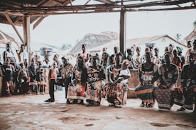 A community gathering scene with a group of people seated and standing under a rustic shelter. The people are dressed in vibrant, colorful clothing with intricate patterns. Some women have baskets balanced on their heads. In the background, traditional thatched-roof houses are visible, as well as several children standing and watching.