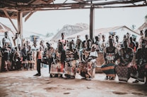 A community gathering scene with a group of people seated and standing under a rustic shelter. The people are dressed in vibrant, colorful clothing with intricate patterns. Some women have baskets balanced on their heads. In the background, traditional thatched-roof houses are visible, as well as several children standing and watching.