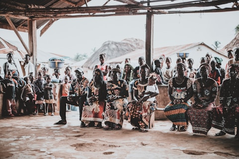 A community gathering scene with a group of people seated and standing under a rustic shelter. The people are dressed in vibrant, colorful clothing with intricate patterns. Some women have baskets balanced on their heads. In the background, traditional thatched-roof houses are visible, as well as several children standing and watching.