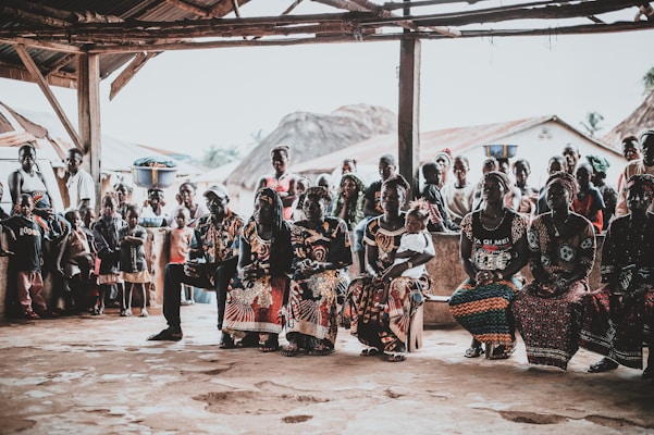 A community gathering scene with a group of people seated and standing under a rustic shelter. The people are dressed in vibrant, colorful clothing with intricate patterns. Some women have baskets balanced on their heads. In the background, traditional thatched-roof houses are visible, as well as several children standing and watching.