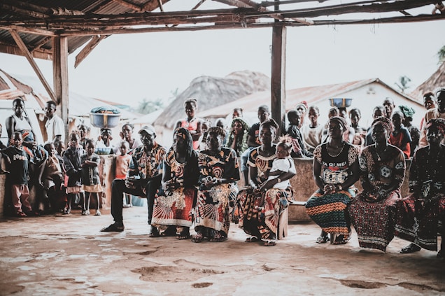 A community gathering scene with a group of people seated and standing under a rustic shelter. The people are dressed in vibrant, colorful clothing with intricate patterns. Some women have baskets balanced on their heads. In the background, traditional thatched-roof houses are visible, as well as several children standing and watching.