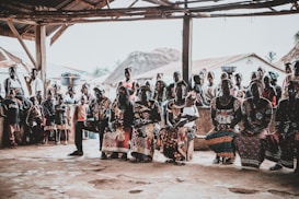 A community gathering scene with a group of people seated and standing under a rustic shelter. The people are dressed in vibrant, colorful clothing with intricate patterns. Some women have baskets balanced on their heads. In the background, traditional thatched-roof houses are visible, as well as several children standing and watching.