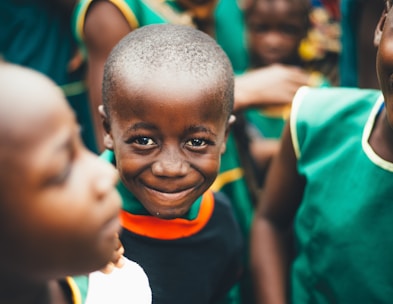 A smiling child is surrounded by other children wearing green clothing. The focus is on the child in the center who is looking directly at the camera with a joyful expression. The background is slightly blurred, emphasizing the central figure.