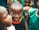 A smiling child is surrounded by other children wearing green clothing. The focus is on the child in the center who is looking directly at the camera with a joyful expression. The background is slightly blurred, emphasizing the central figure.
