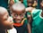 A smiling child is surrounded by other children wearing green clothing. The focus is on the child in the center who is looking directly at the camera with a joyful expression. The background is slightly blurred, emphasizing the central figure.