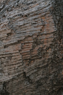 A textured rock face with layers of stacked stone displaying shades of gray and brown, with elements of weathering and erosion visible.
