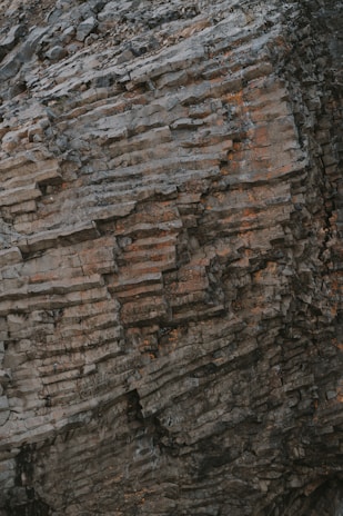 A textured rock face with layers of stacked stone displaying shades of gray and brown, with elements of weathering and erosion visible.