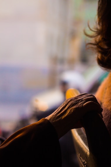 A close-up of an elderly hand resting on the back of a chair, with warm lighting casting shadows and highlighting the details of the skin. The background is blurred, suggesting a soft focus on the subject.