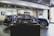 A showroom interior featuring a sleek, blue car prominently displayed, with a smaller, detailed model of the same car encased in glass at the forefront. The environment has a polished and modern aesthetic, with tiled flooring, bright lighting, and a television screen in the background.