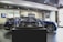 A showroom interior featuring a sleek, blue car prominently displayed, with a smaller, detailed model of the same car encased in glass at the forefront. The environment has a polished and modern aesthetic, with tiled flooring, bright lighting, and a television screen in the background.