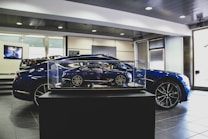 A showroom interior featuring a sleek, blue car prominently displayed, with a smaller, detailed model of the same car encased in glass at the forefront. The environment has a polished and modern aesthetic, with tiled flooring, bright lighting, and a television screen in the background.