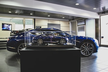 A showroom interior featuring a sleek, blue car prominently displayed, with a smaller, detailed model of the same car encased in glass at the forefront. The environment has a polished and modern aesthetic, with tiled flooring, bright lighting, and a television screen in the background.