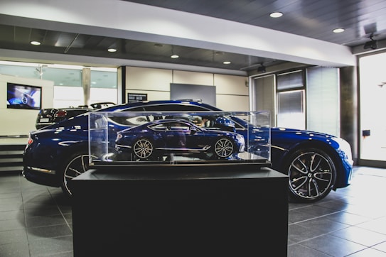 A showroom interior featuring a sleek, blue car prominently displayed, with a smaller, detailed model of the same car encased in glass at the forefront. The environment has a polished and modern aesthetic, with tiled flooring, bright lighting, and a television screen in the background.