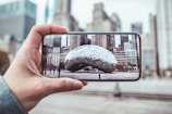A person holds a smartphone capturing an image of a large reflective sculpture in an urban plaza. The background shows tall buildings in a cityscape with a mix of modern and classical architectural designs.