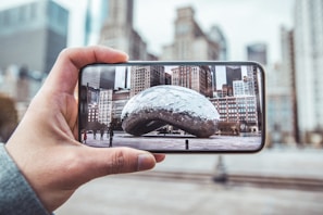 A person holds a smartphone capturing an image of a large reflective sculpture in an urban plaza. The background shows tall buildings in a cityscape with a mix of modern and classical architectural designs.