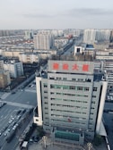 An urban landscape with a high-rise building in the foreground, featuring a sign with large red characters on the roof. The view includes several other tall buildings stretching into the distance, indicating a densely built area. The streets are wide with visible road markings and light traffic.