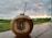 A coconut with a straw inserted sits on a wooden surface. The coconut is branded with the words 'Empire Fit Club Bali'. In the background, there is a lush green field and an overcast sky with orange-yellow tinges from a setting or rising sun.