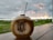 A coconut with a straw inserted sits on a wooden surface. The coconut is branded with the words 'Empire Fit Club Bali'. In the background, there is a lush green field and an overcast sky with orange-yellow tinges from a setting or rising sun.
